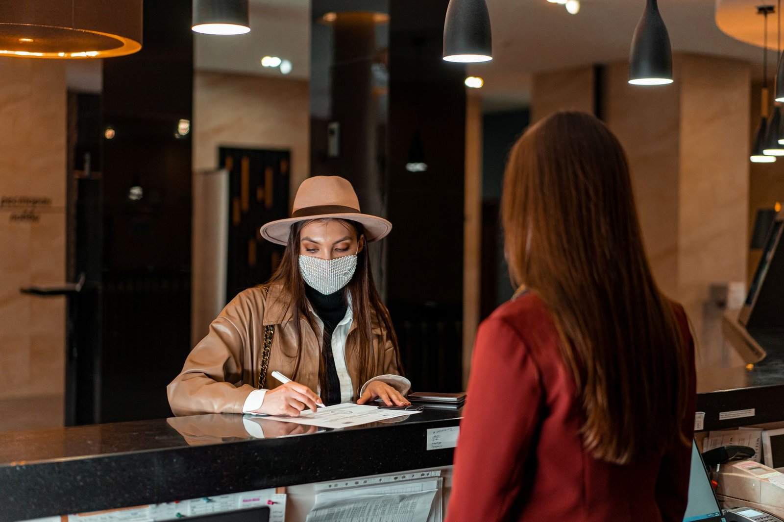 A woman in a hat and face mask checks in at a hotel reception, interacting with a receptionist